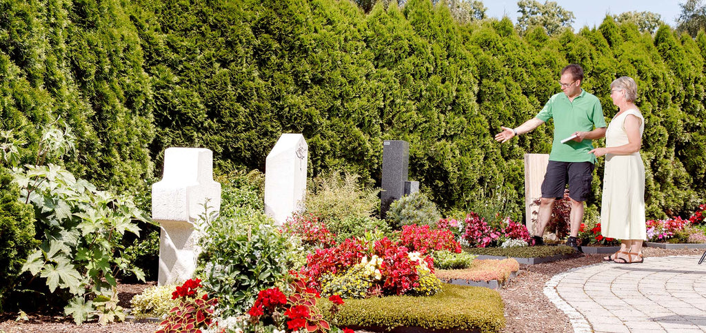 Geschäftsführer Arnold Konzelmann berät Kundin bei der Grabbepflanzung und Grabpflege. Sie stehen auf dem Friedhof vor gepflegten Gräbern.