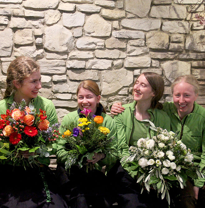 Floristinnen und Gärtnerinnen freudig bei der Arbeit. Sie sitzen nebeneinander mit Blumensträuße.